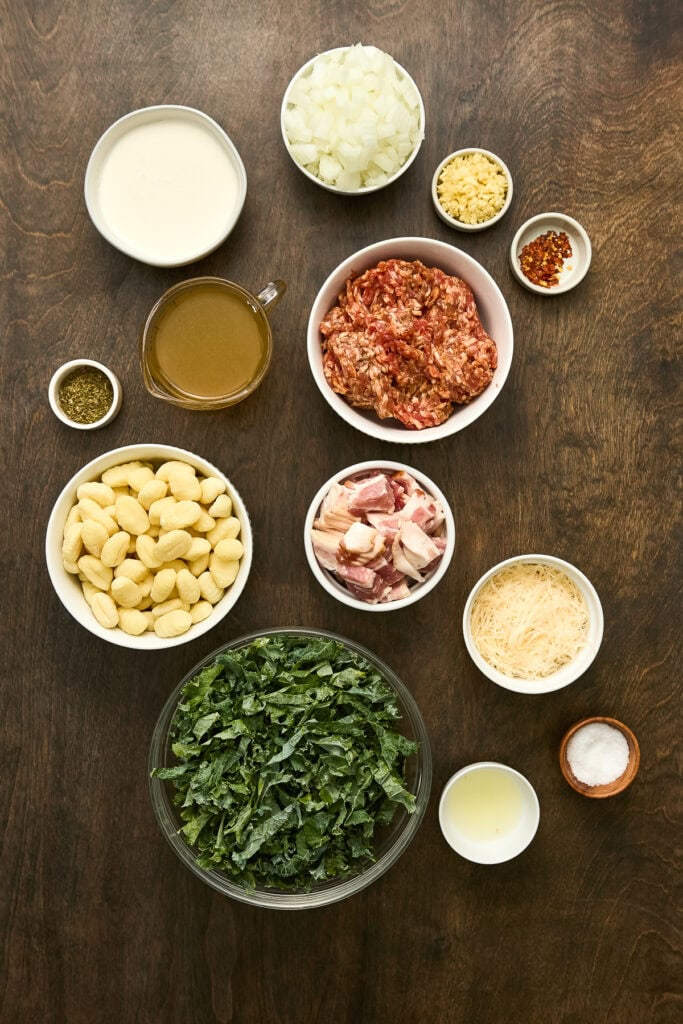 Assorted ingredients for Zuppa Toscana Gnocchi including ground meat, kale, onions, garlic, cream, and seasonings arranged on a wooden surface.