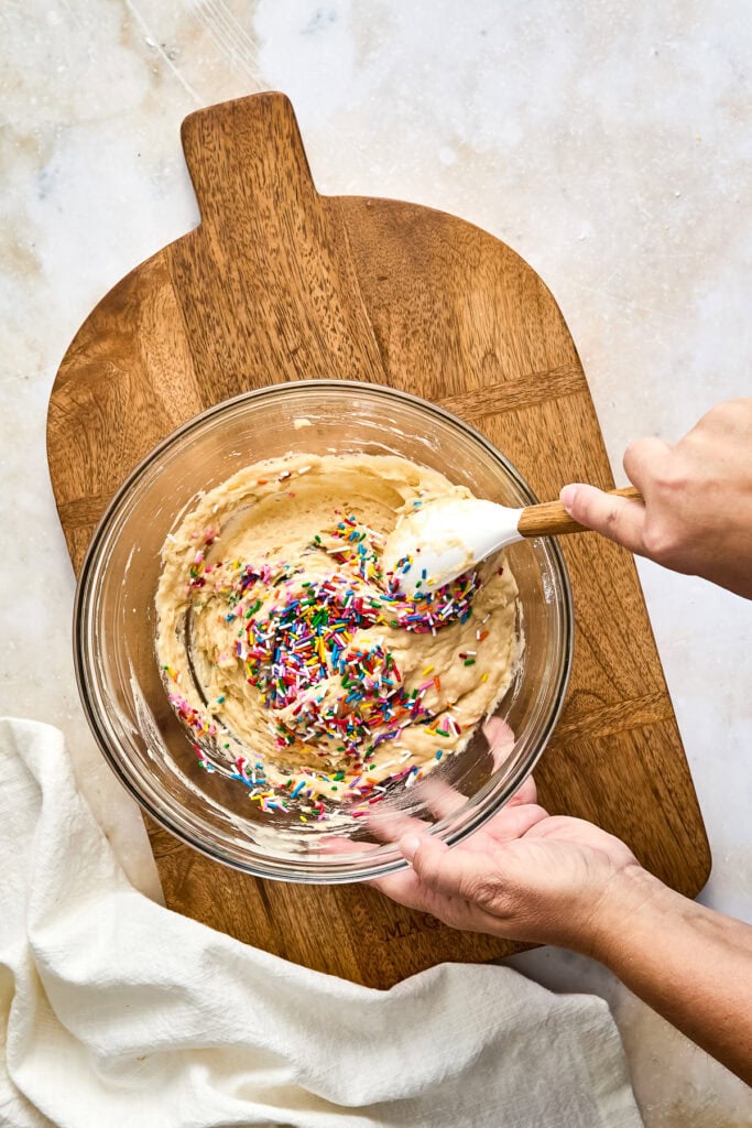 Close-up of a glass mixing bowl with muffin batter and colorful sprinkles, being stirred with a spatula on a wooden cutting board.