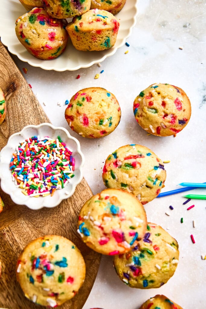 Freshly baked mini muffins colorful sprinkles on a white surface, with a bowl of rainbow sprinkles and scattered sprinkles around.