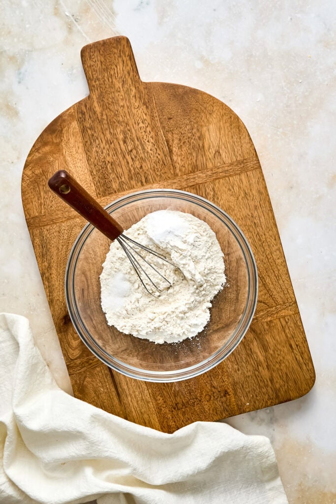 Glass bowl with flour and a whisk on a wooden cutting board, ready for baking, with a white cloth nearby.