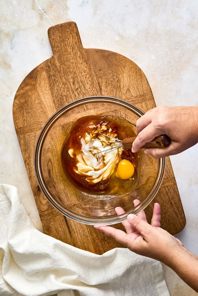 Person mixing cake batter with eggs, greek yogurt, and maple syrup in a glass bowl on a wooden board.