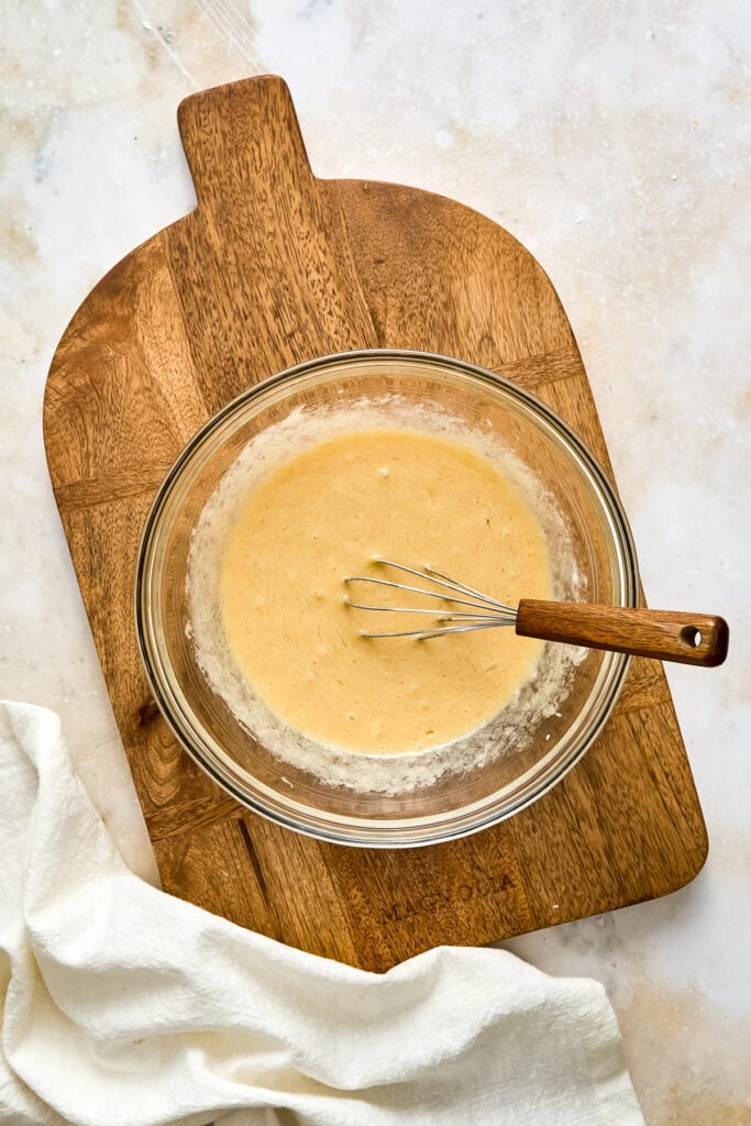 Creamy muffin batter in a glass bowl with a whisk and wooden spatula on a wooden cutting board, ready for baking.