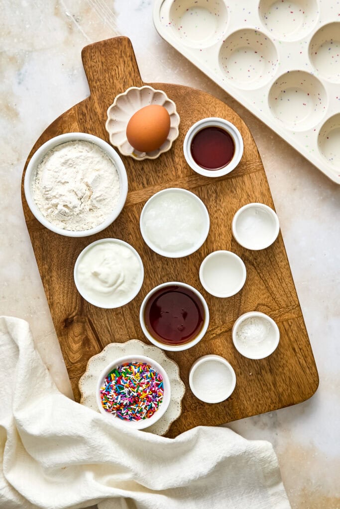 Assorted baking ingredients and supplies on a wooden cutting board, including flour, eggs, sprinkles, and various liquids, ready for baking.