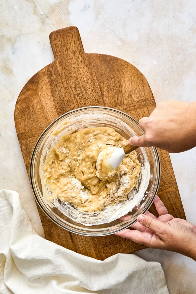 Person mixing muffin batter in a glass bowl on a wooden cutting board, preparing for baking at Stuck On Sweet.