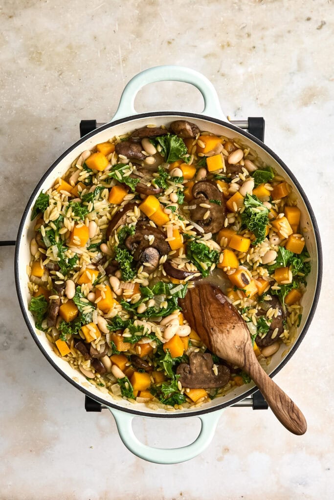 A skillet of creamy butternut squash orzo with mushrooms, kale, and herbs, served in a white pan with a wooden spoon, on a neutral background.