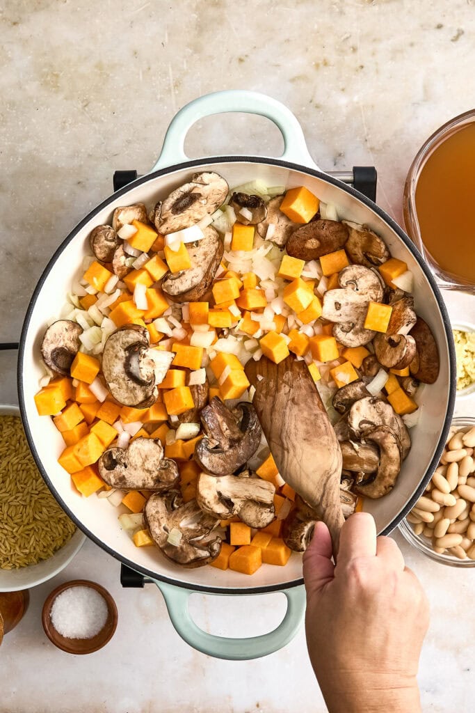 Close-up of a skillet with cooked butternut squash, mushrooms, and kale, ready for serving, with a hand stirring the ingredients.