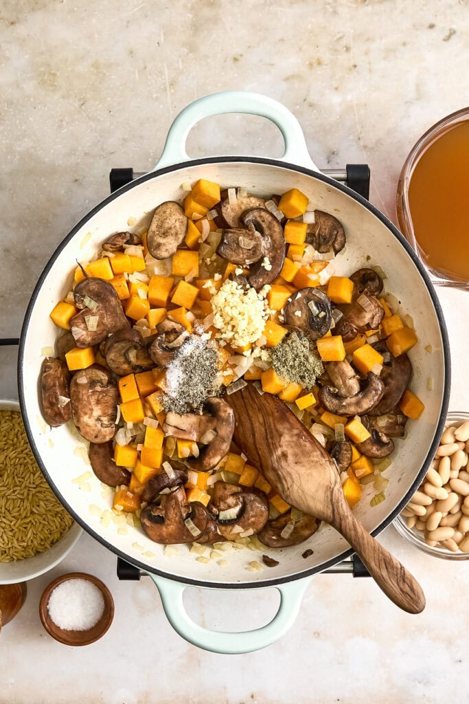 A skillet of creamy butternut squash orzo with mushrooms, diced butternut squash, garlic, and herbs, served with a side of broth and beans.