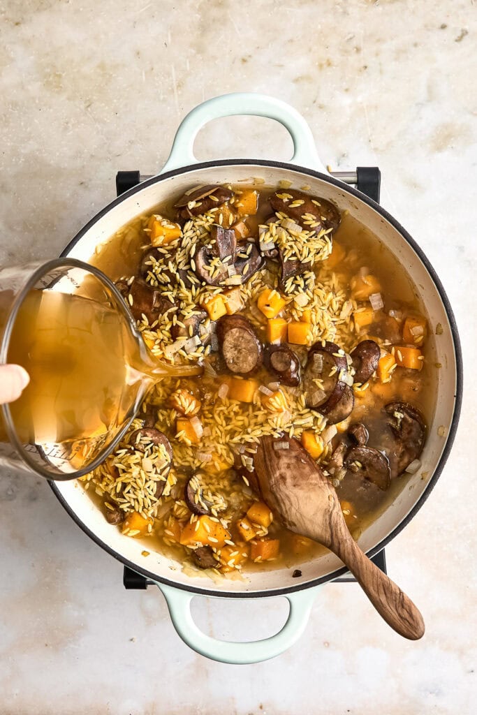 A pot of creamy butternut squash orzo with mushrooms, carrots, and kale being cooked on a stovetop, with broth being poured in.