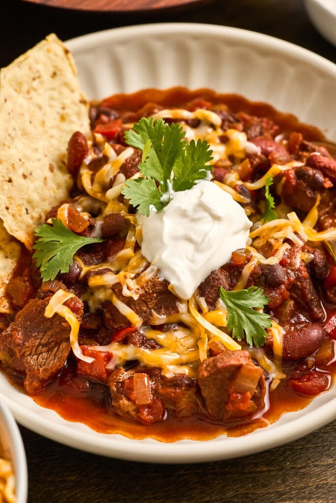 A bowl of hearty chuck roast chili topped with shredded cheese, sour cream, cilantro, and a side of cornbread, perfect for comfort food lovers.