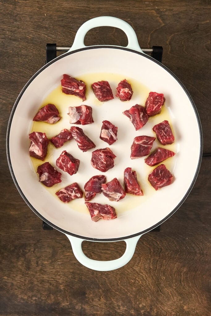Raw beef chunks cooking in a white pot with oil, on a wooden surface, ready for making beef stew.