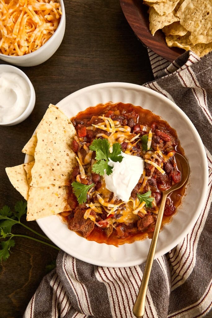 A bowl of hearty chuck roast chili topped with shredded cheese, sour cream, and cilantro, served with tortilla chips and side dishes on a dark wooden table.