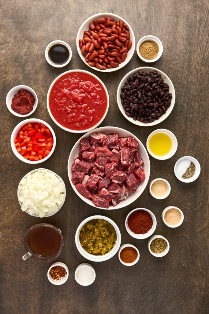 Assorted ingredients for beef chili including beans, tomatoes, beef, spices, and seasonings arranged on a wooden surface.
