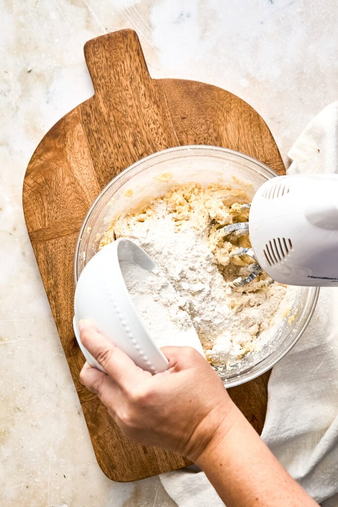 Person mixing dough in a glass bowl using a hand mixer on a wooden cutting board, preparing sweet baked goods.