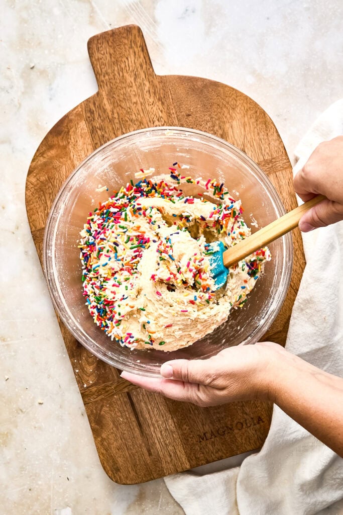A person mixing colorful sprinkles into a creamy cake batter in a glass bowl on a wooden cutting board.