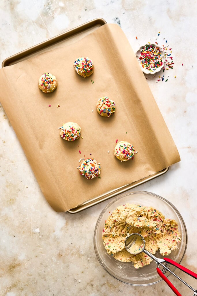 Fresh cookie dough with colorful sprinkles and a scoop, ready for baking, on a baking sheet with a single decorated cookie on the side.