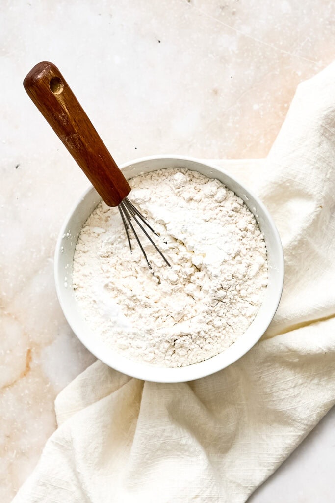 Close-up of a white bowl filled with flour, with a wooden-handled whisk resting inside, on a light surface with a white cloth.