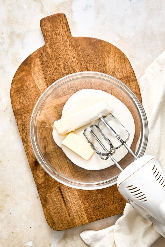 Cream cheese and butter in a glass bowl with an electric mixer on a wooden cutting board, ready for baking or dessert preparation.