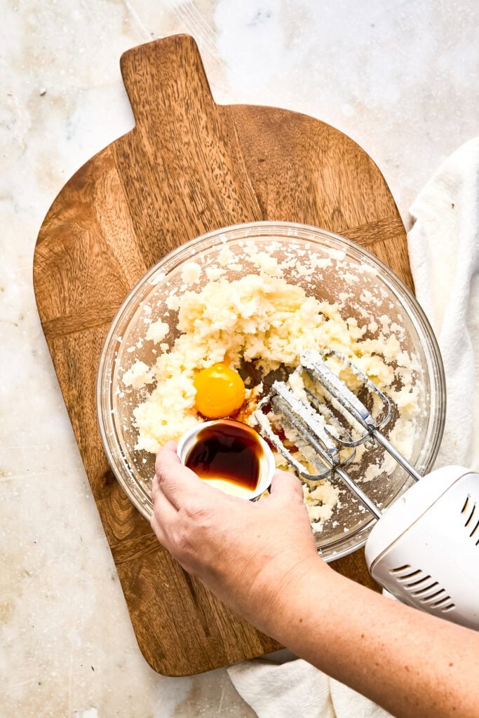Person mixing cookie dough with an electric mixer, adding vanilla extract in a glass bowl on a wooden cutting board.