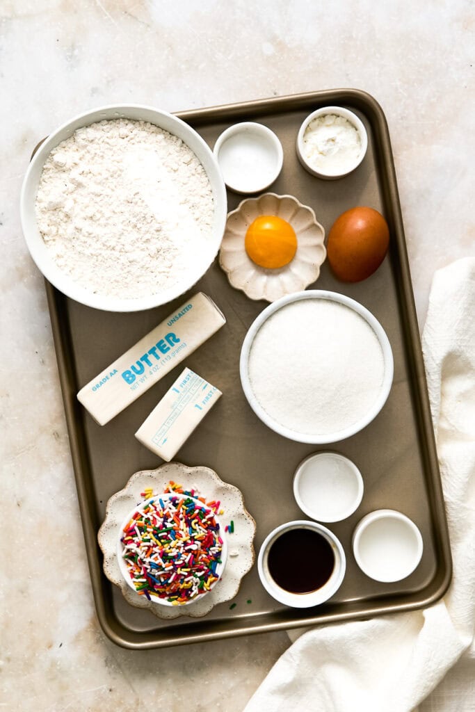 Assorted baking ingredients on a tray including flour, eggs, butter, sugar, vanilla, and sprinkles for baking delicious desserts.
