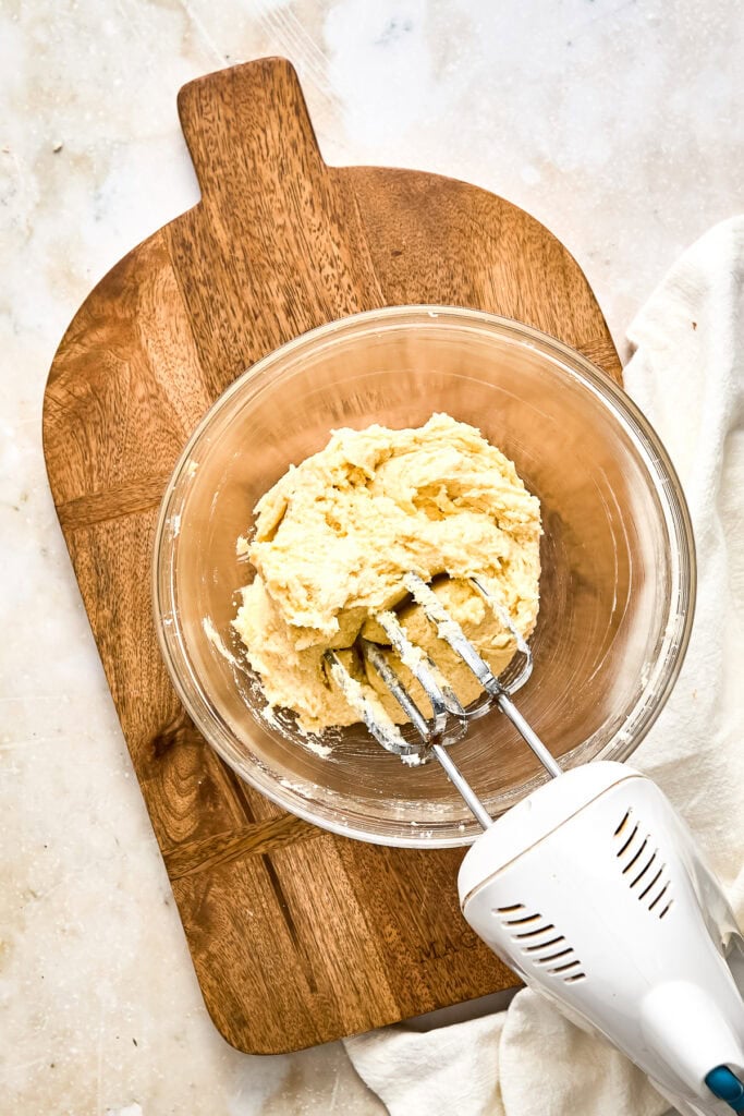 Close-up of cookie dough in a glass bowl with a hand mixer on a wooden cutting board, ready for baking.