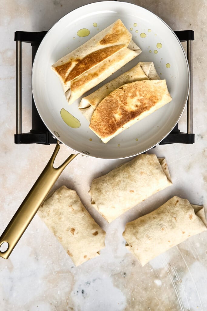 Freshly cooked chicken burritos in a white skillet with golden-brown tortillas, ready to serve, on a light marble surface.