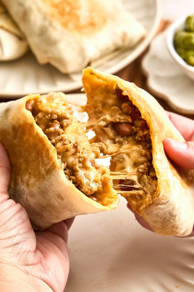 Close-up of a freshly baked beef empanada being broken in half, revealing savory meat filling inside, served with side dishes in the background.