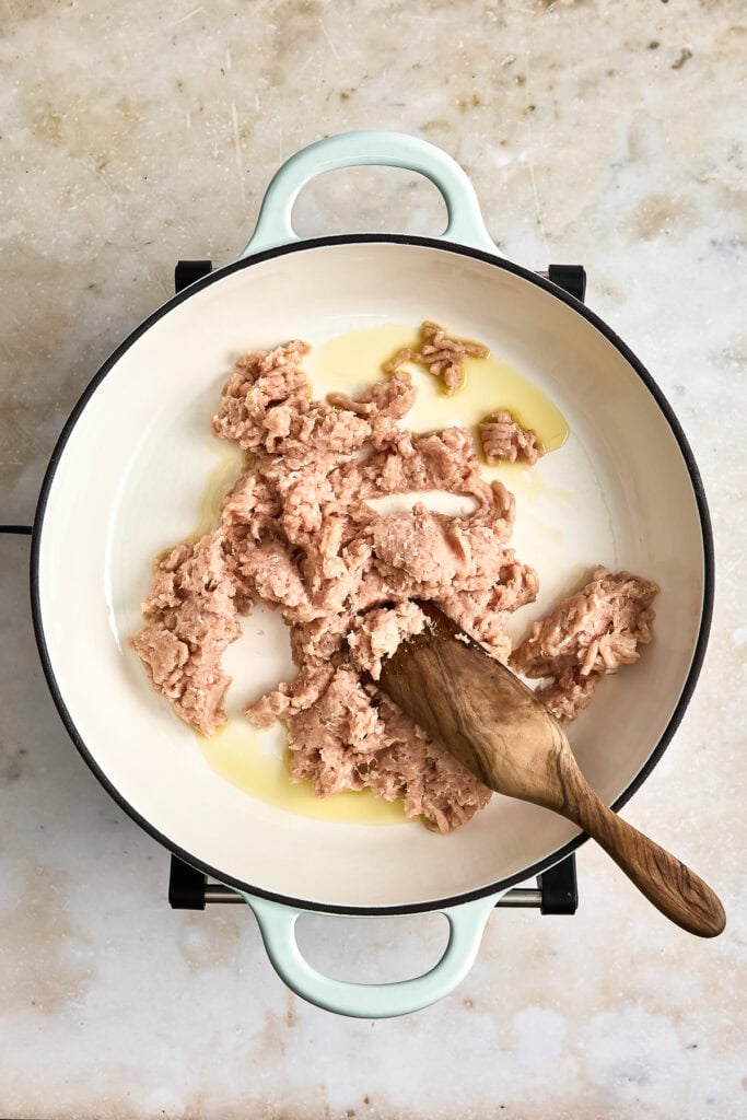 Ground meat being cooked in a white skillet with a wooden spoon, with oil or butter visible, on a light-colored surface.
