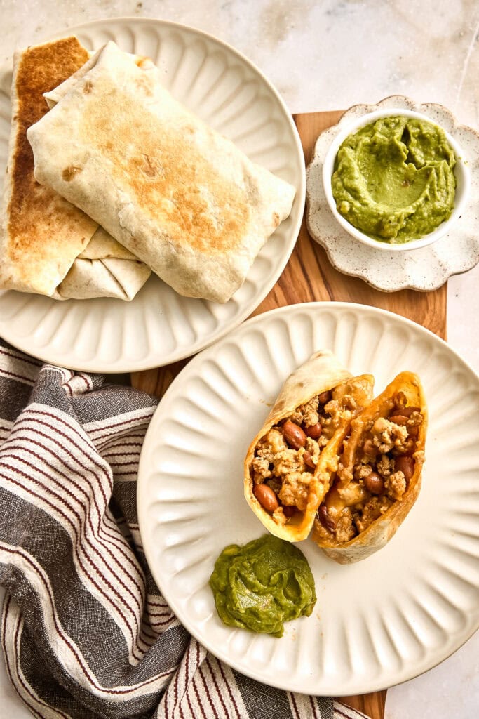 Freshly made chicken burritos filled with seasoned meat and beans, served with a side of vibrant green guacamole on a white plate.