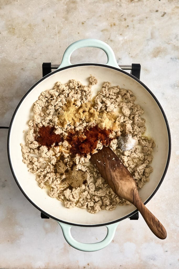 Ground chicken being cooked with spices in a white skillet, with a wooden spoon stirring the mixture, on a light textured surface.