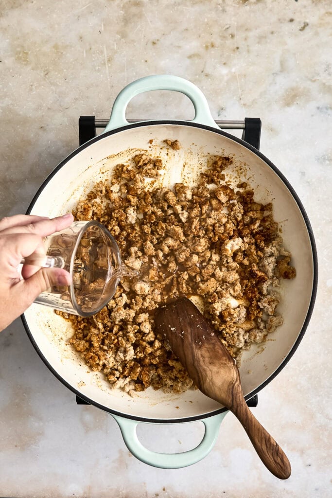 ground chicken and spiced in a white skillet with a wooden spoon, with a hand pouring in liquid, on a light textured surface.