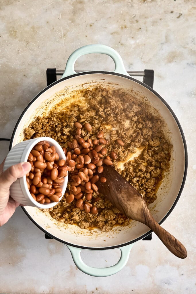 A person adding cooked beans into a pot with ground chicken and onions, cooking on a stovetop, with a wooden spoon inside the pot.