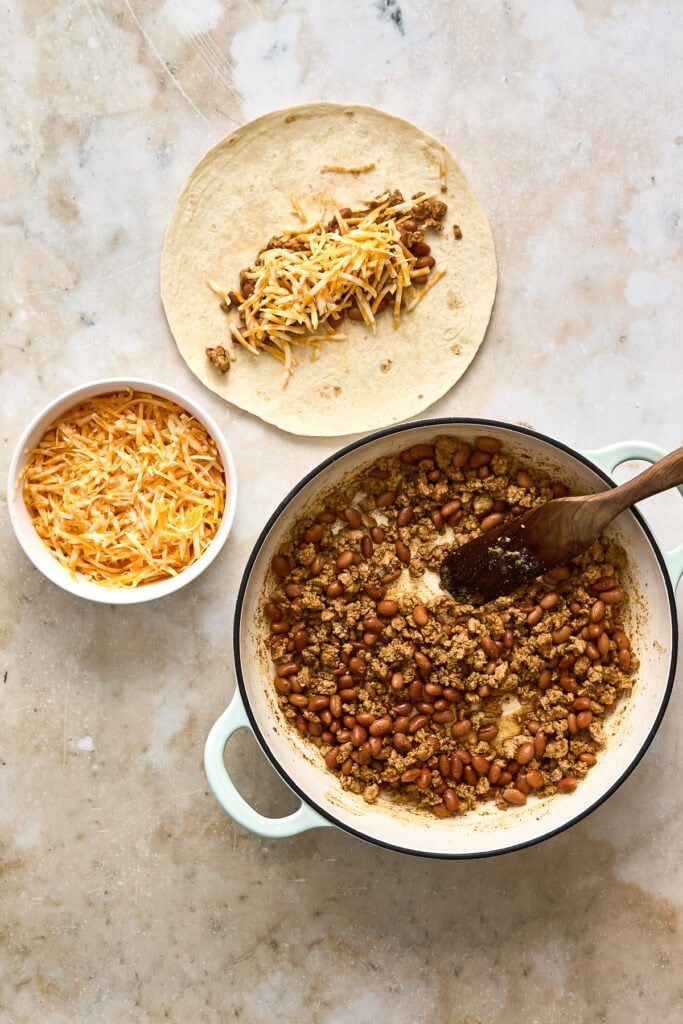 Close-up of chicken and bean filling cooking in a pot, with a taco shell filled with shredded cheese and seasoned meat and beans on a plate.