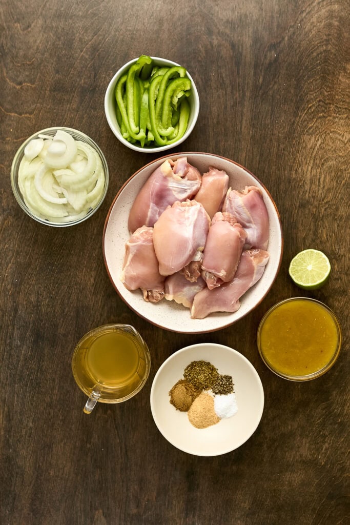 Raw chicken thighs, sliced green bell peppers, sliced onions, lime, and spices on a wooden surface for fajita preparation.