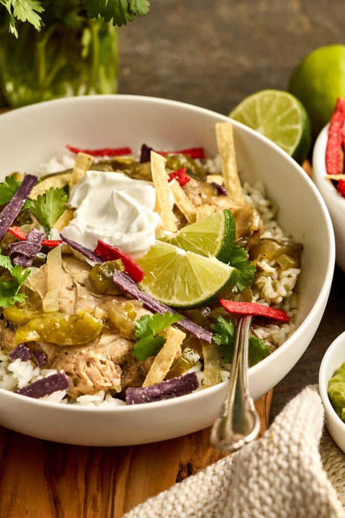 A bowl of salsa verde chicken over rice garnished with lime wedges, cilantro, sour cream, and crispy tortilla strips, served on a rustic wooden table.