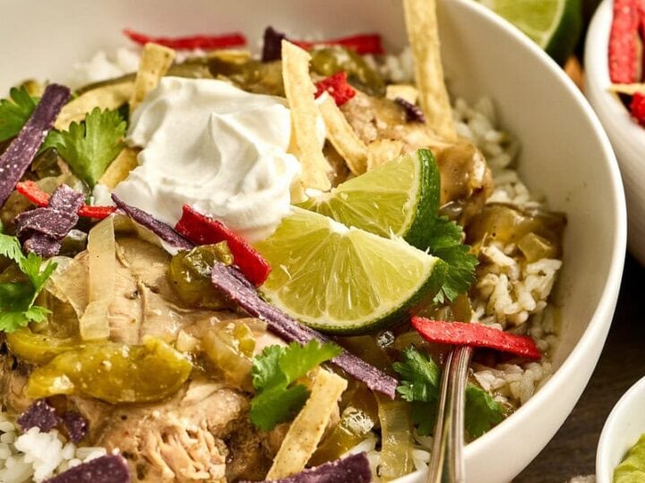 A bowl of salsa verde chicken over rice garnished with lime wedges, cilantro, sour cream, and crispy tortilla strips, served on a rustic wooden table.
