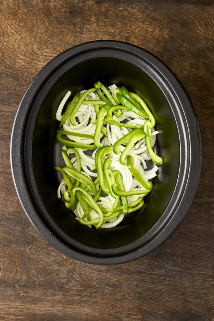 Sliced green bell peppers in a black slow cooker on a wooden surface, ready for cooking or seasoning.