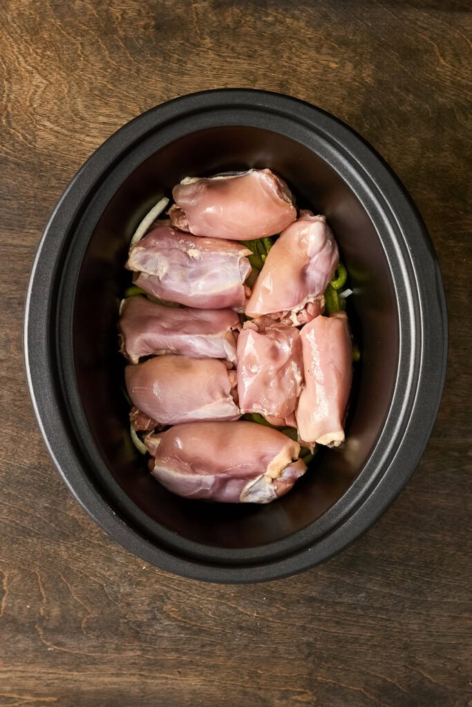 Raw chicken thighs placed in a black slow cooker, ready for cooking, on a wooden surface.