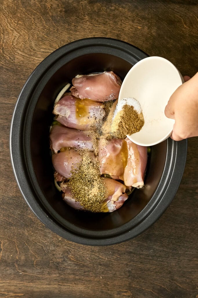 Raw chicken pieces in a black slow cooker with herbs and spices, ready for cooking.