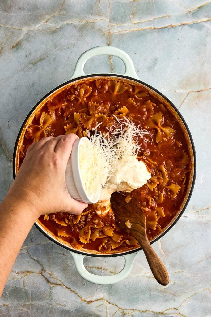 parmesan cheese, ricotta cheese and balsamic vinegar being added to a skillet with beef tomato sauce and bowtie pasta.