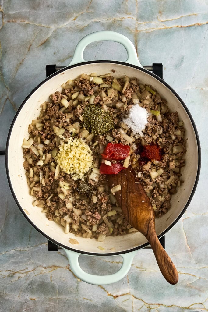 Ground beef and dice onion cooking in a white skillet with herbs, garlic, salt, and pepper, tomato paste on a marble countertop.