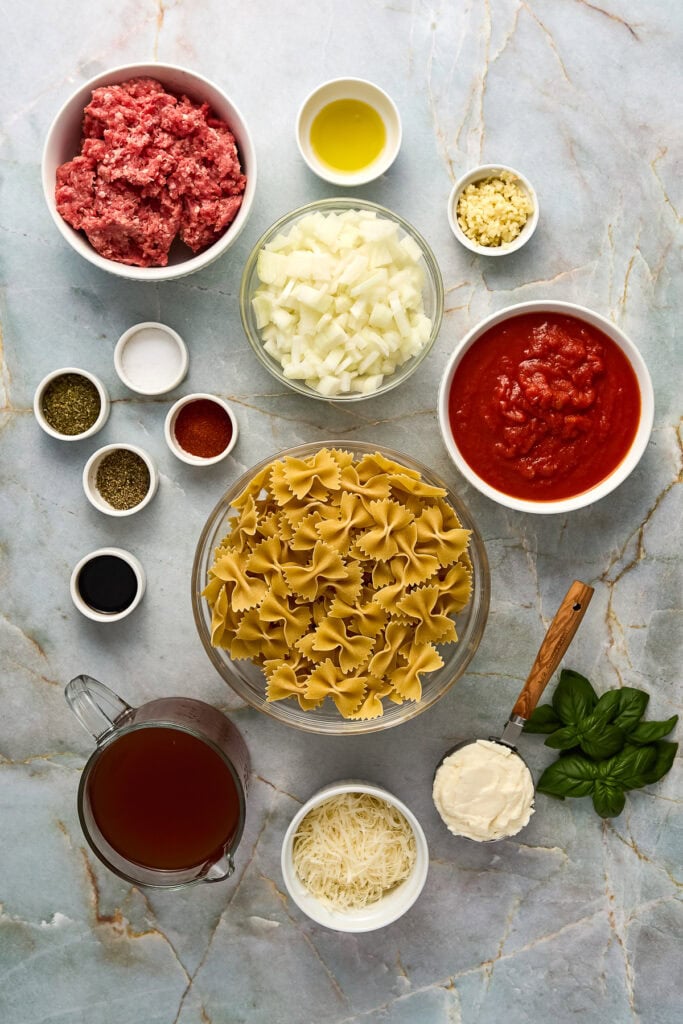 Assorted ingredients for making homemade creamy tomato ricotta pasta with meat sauce, including ground meat, pasta, onions, tomato sauce, Parmesan cheese, ricotta cheese and herbs.
