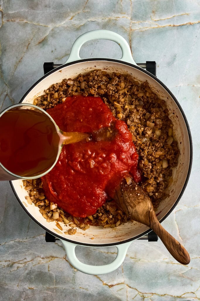 tomato sauce and beef stock being poured into a skillet with cooked ground beef, spices and herbs.