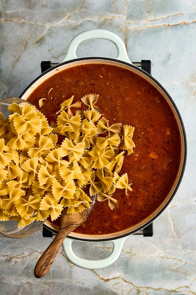 Uncooked farfalle pasta being added to a simmering pot of meat tomato sauce on a marble countertop.