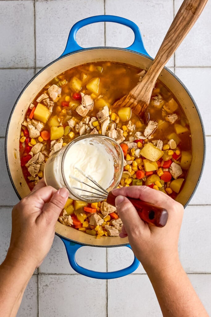 Person adding heavy cream to a pot of homemade chicken corn chowder soup with carrots, potatoes, and corn.
