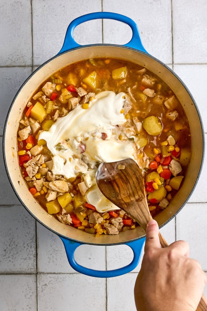 A pot of homemade chicken corn chowder being stirred with a wooden spoon, featuring diced potatoes, carrots, and chicken in a savory broth.