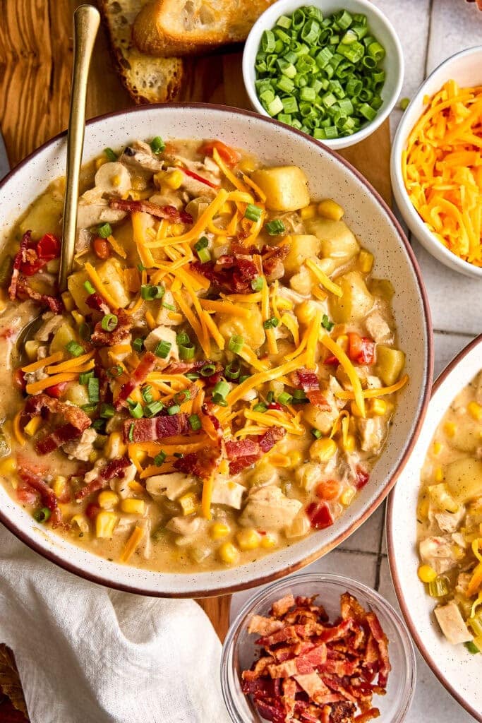 A hearty bowl of creamy chicken and corn chowder topped with shredded cheese, chopped green onions, and crispy bacon, served with bread and fresh toppings.