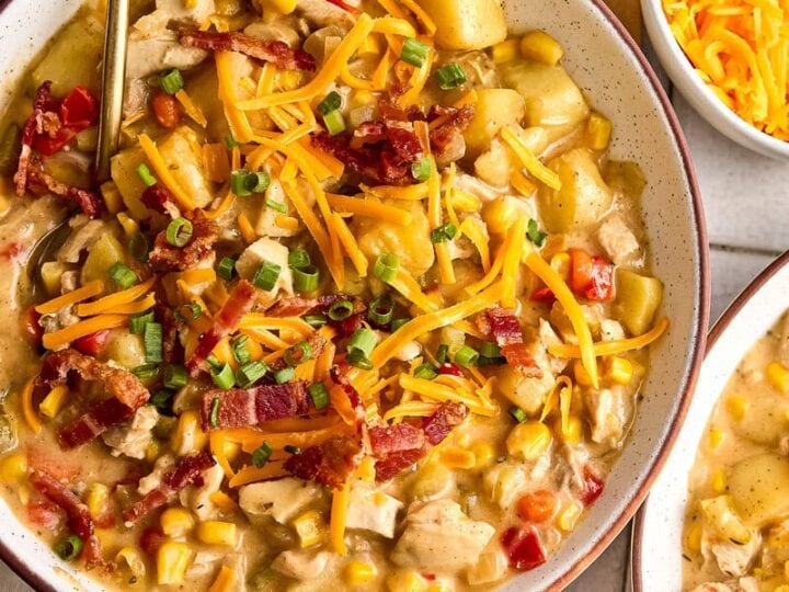 A hearty bowl of creamy chicken and corn chowder topped with shredded cheese, chopped green onions, and crispy bacon, served with bread and fresh toppings.