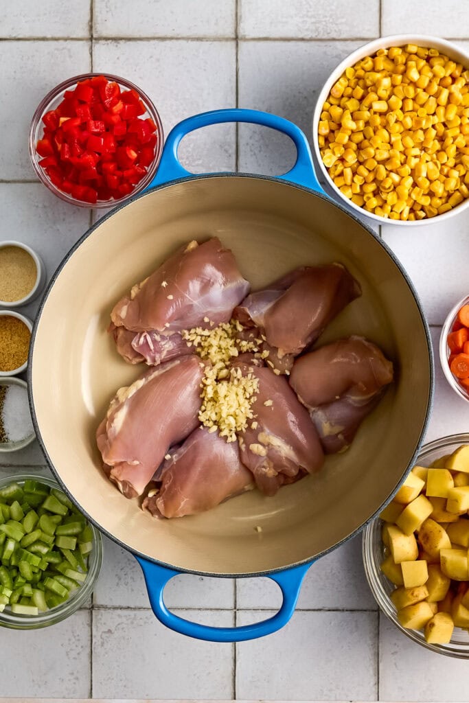 Raw chicken thighs with minced garlic in a blue casserole dish surrounded by chopped vegetables and canned corn for chicken corn chowder