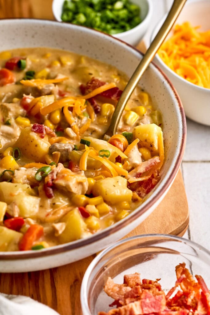 A bowl of hearty chicken and corn chowder soup with corn, potatoes, and bacon, served with side bowls of shredded cheese and chopped green onions.
