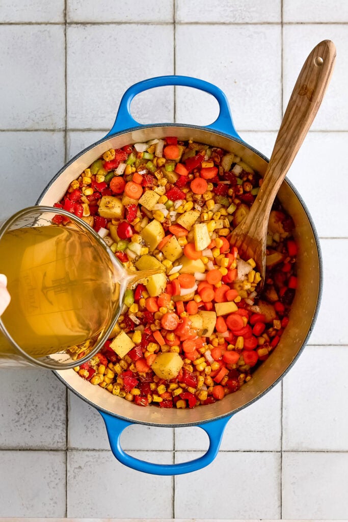 Colorful chicken corn chowder with carrots, corn, and potatoes in a blue pot, with a wooden spoon and a glass of broth, on a white tiled surface.
